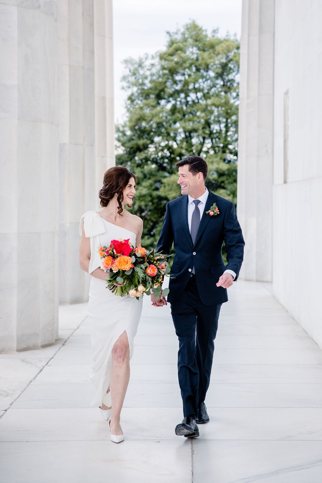 a couple walking hand in hand together on the day of their wedding smiling at each other the woman is holdin ga bouquet of flowers s