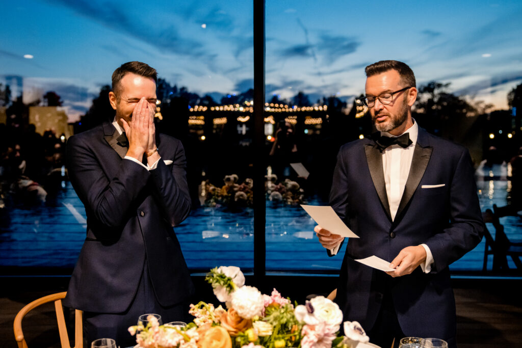 A newlywed giving a speech as they partner stands next to them laughing into their hands 