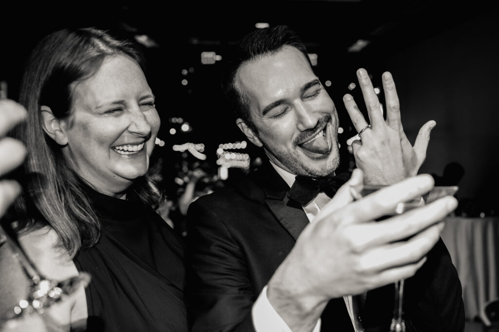 A newlywed with their tongue sticking out showing their wedding ring with someone laughing next to them 