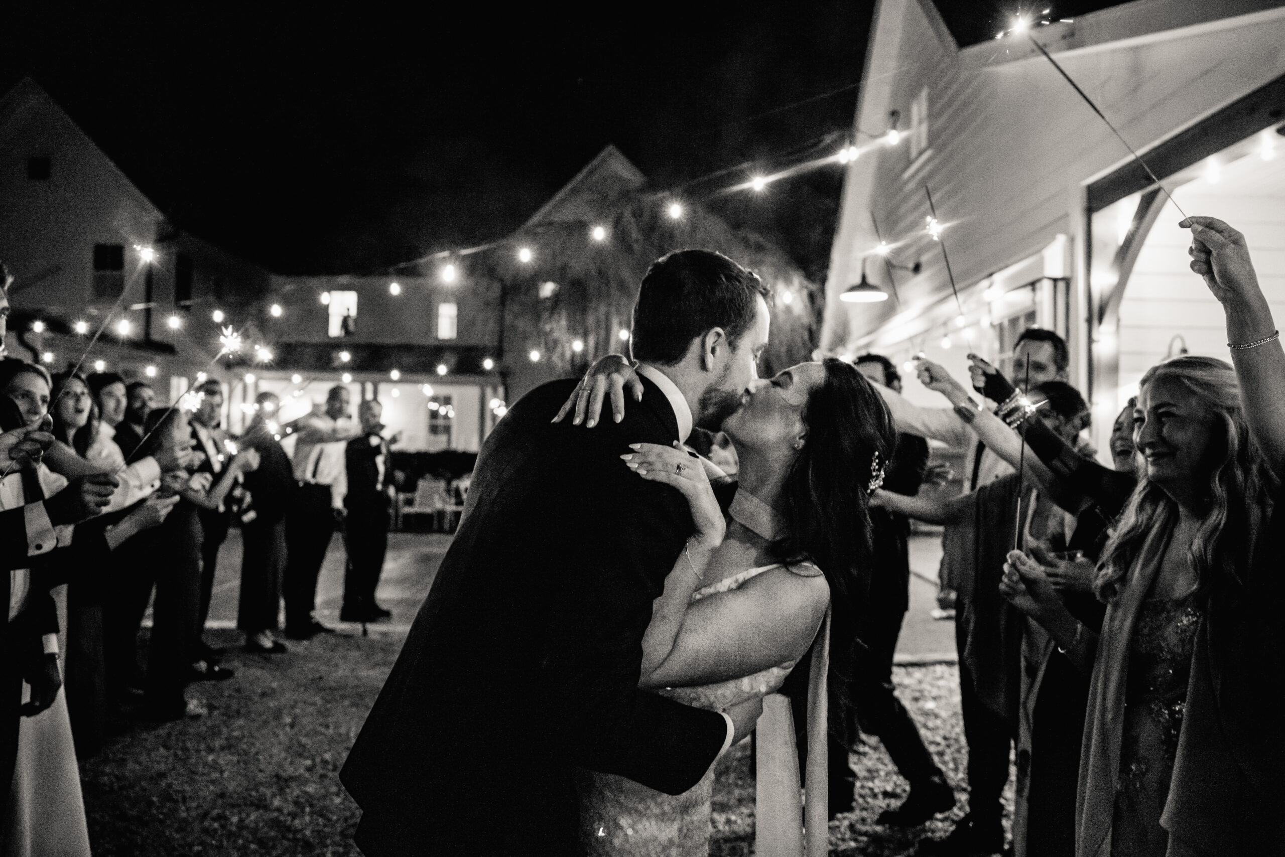 A newlywed couple kissing under twinkle lights as their guests cheer behind them
