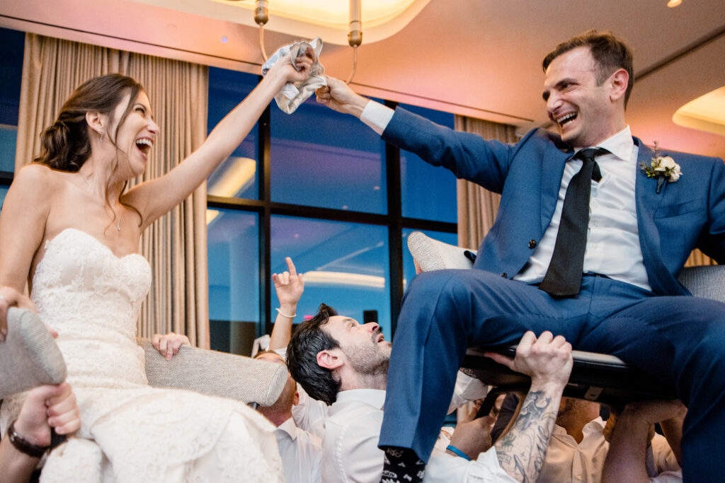 Newlyweds laughing and holding a cloth together as they are held up in chairs 