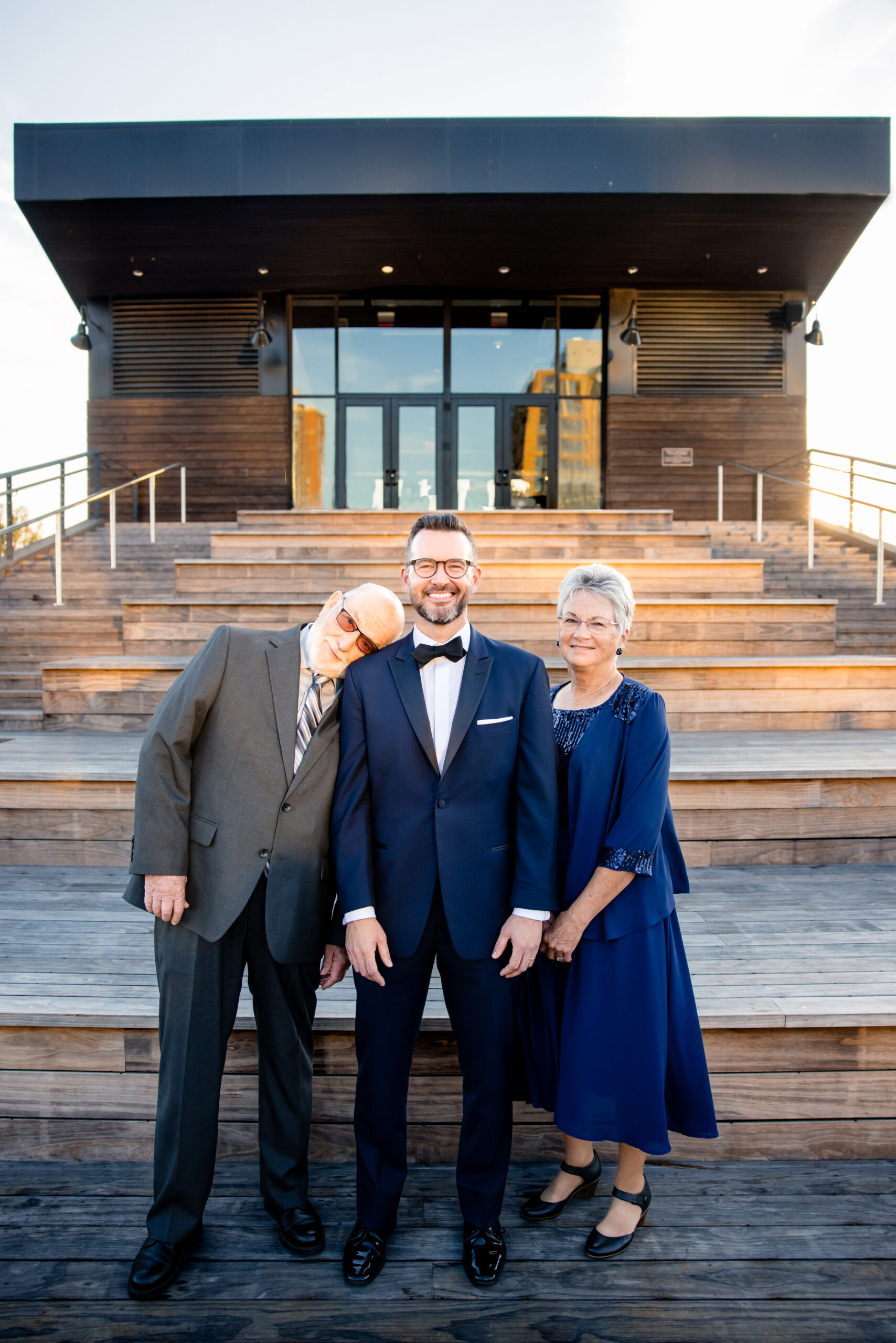 a groom on a set of steps taking a photo with his mom and dad