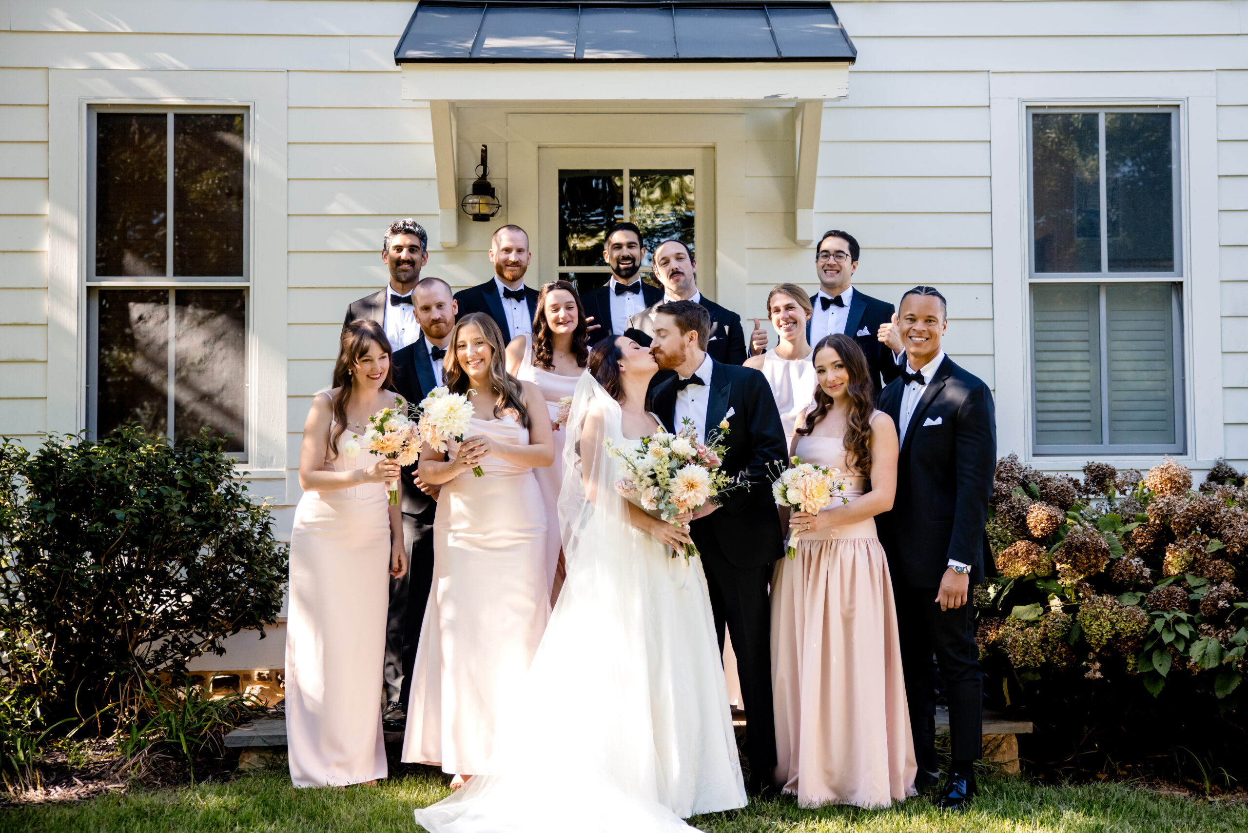 a bride and groom kissing in front of a house taking wedding photos with their wedding party