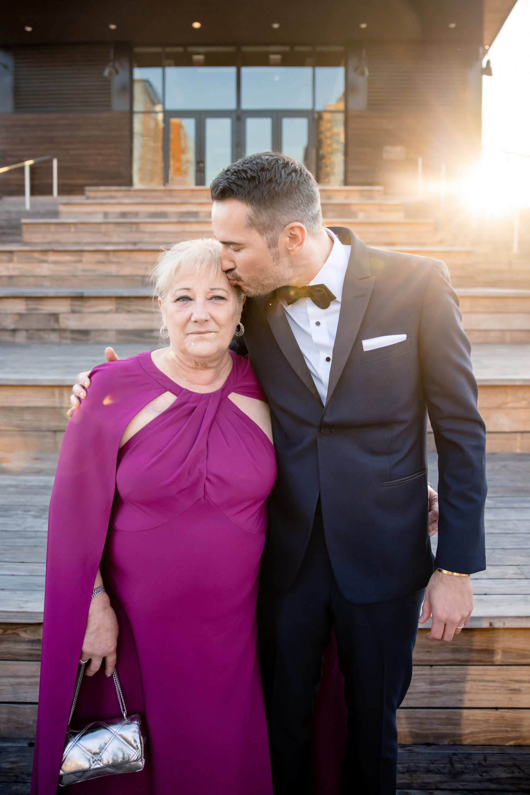 a groom kissing his mother on the side of her forehead with his arm wrapped around her 