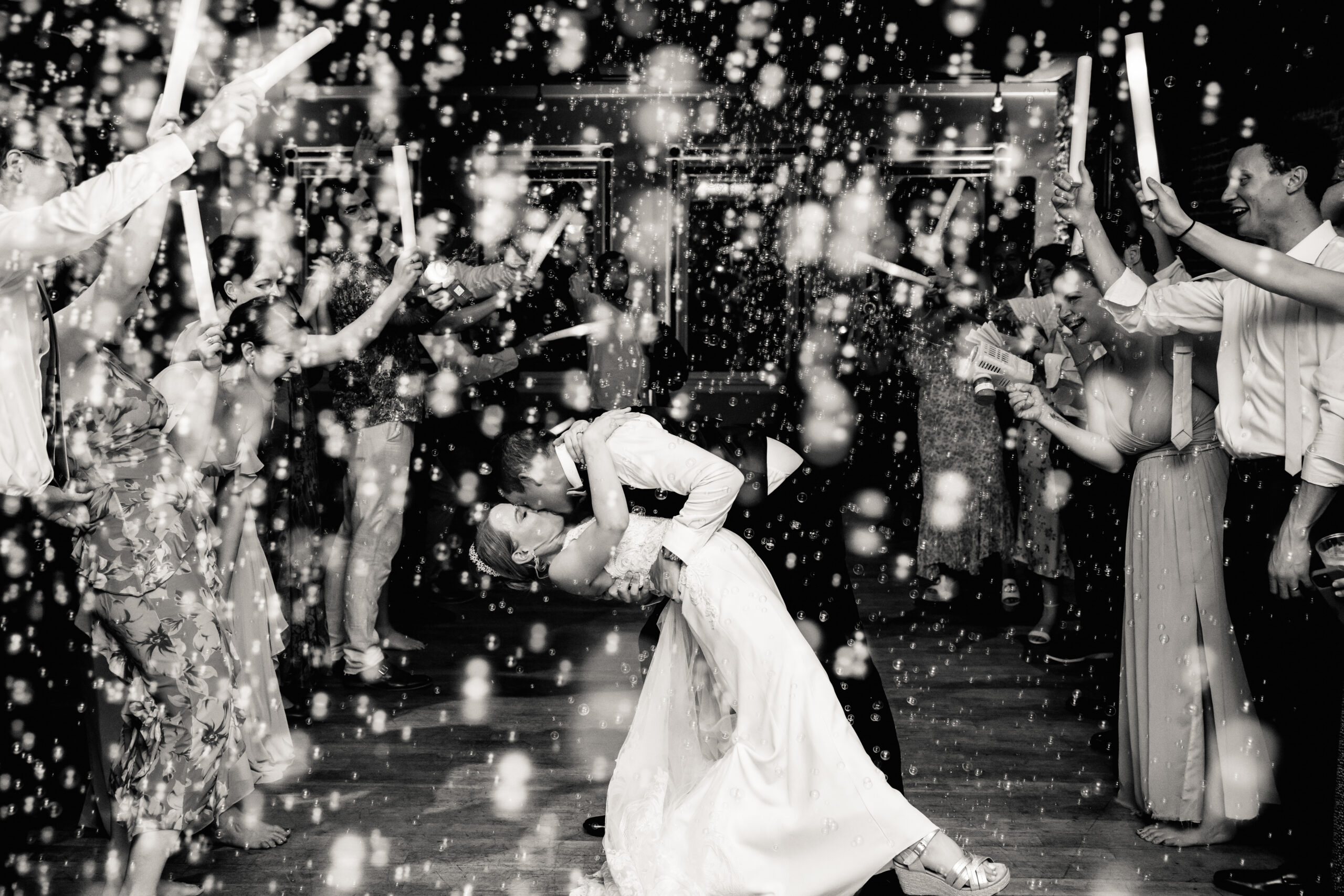 a bride and groom kissing as their guests cheer for them the groom is dipping the bride backwards and the room is filled with bubbles 