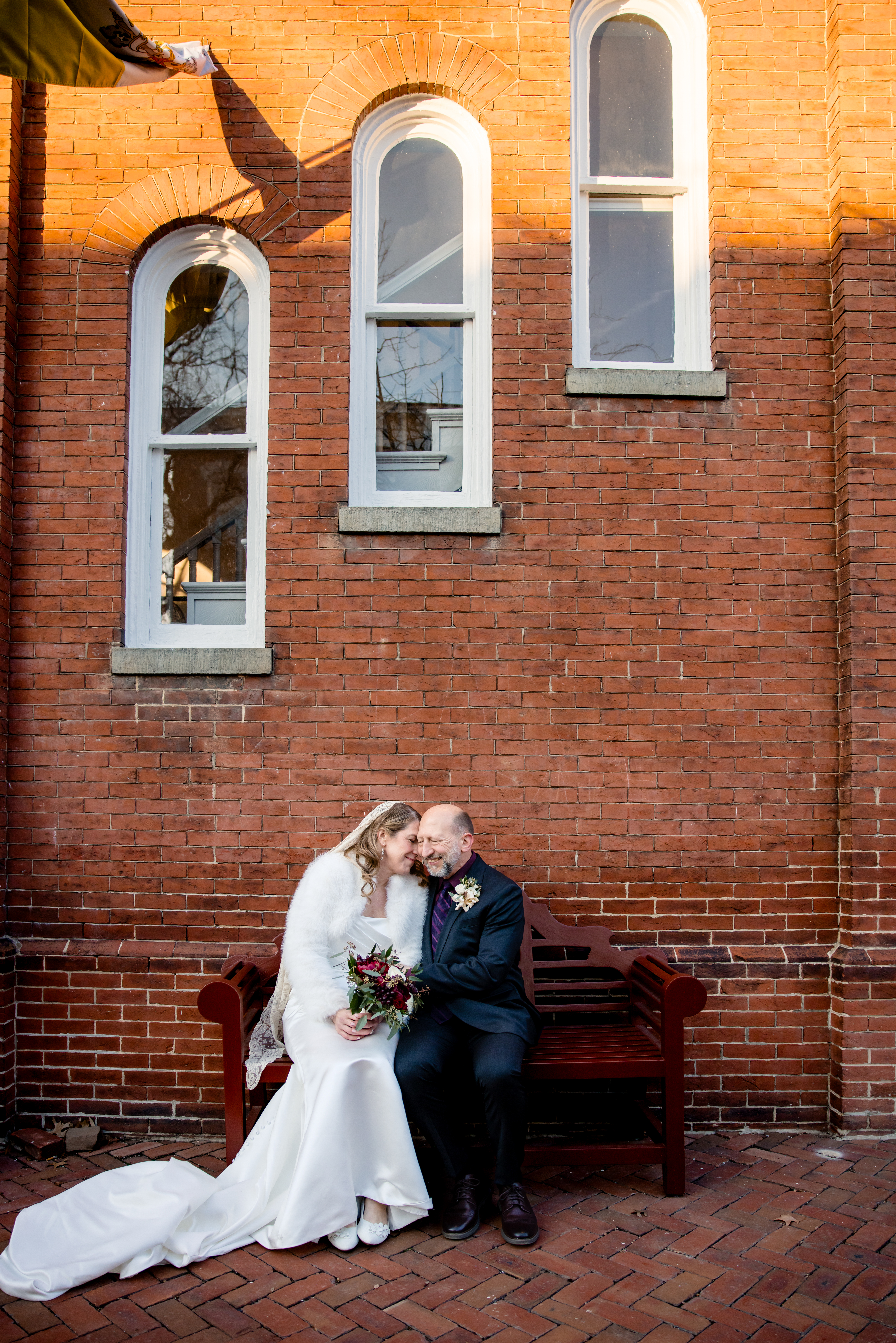 a bride and groom sitting on a bench together leaning towards each other in front of a red brick building 