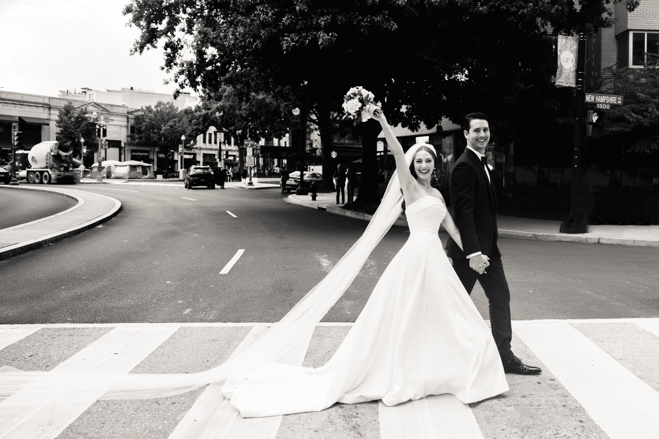 a couple walking hand in hand together on the day of their wedding across a sidewalk 