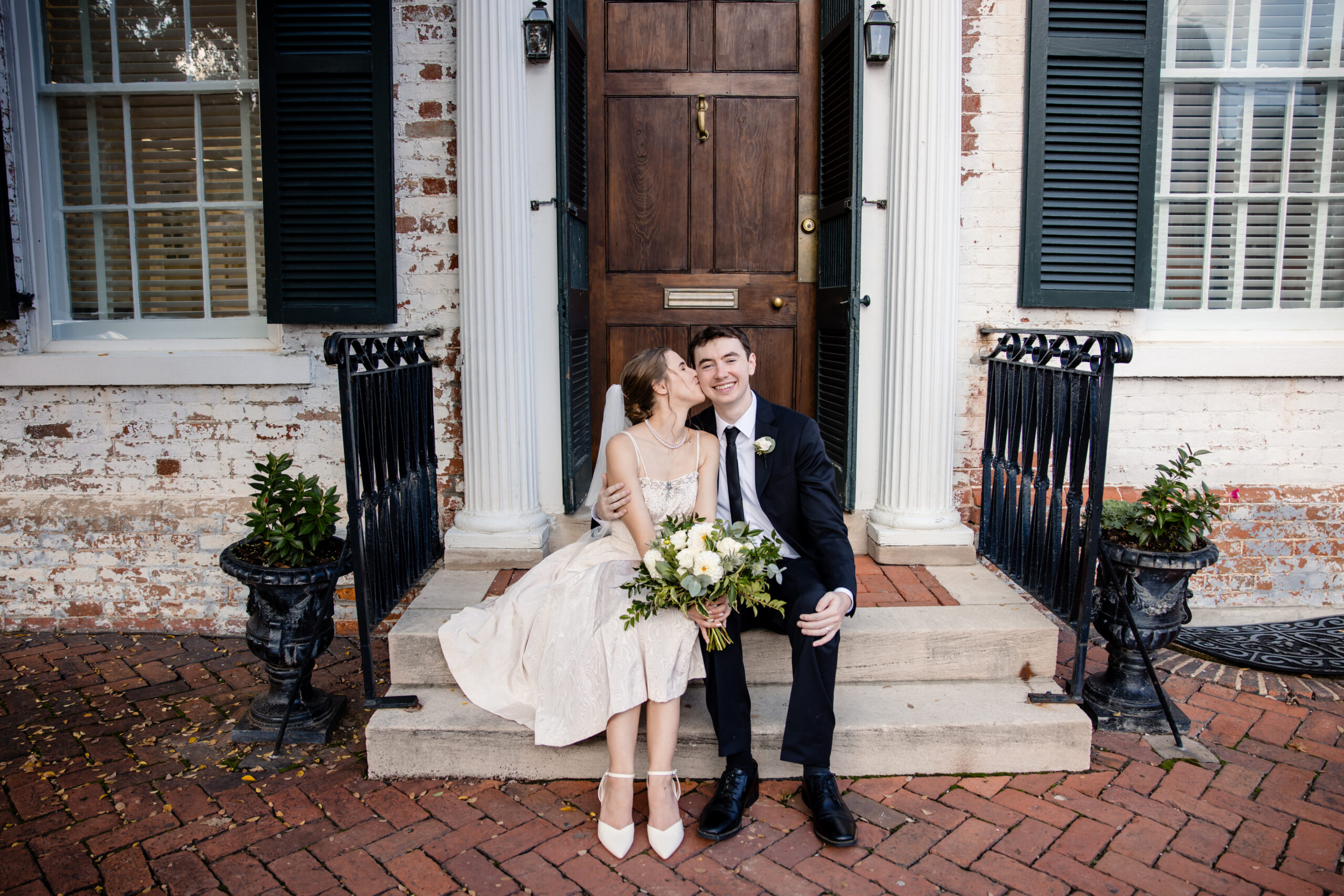 a couple sitting on steps together on the day of their wedding the woman is kissing her partner on his cheek and the man is smiling