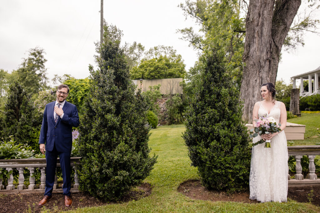 a couple standing side by side together in a garden about to share a first look with each other on the day of their wedding the woman is looking at her partner and the man has his hand on his chest