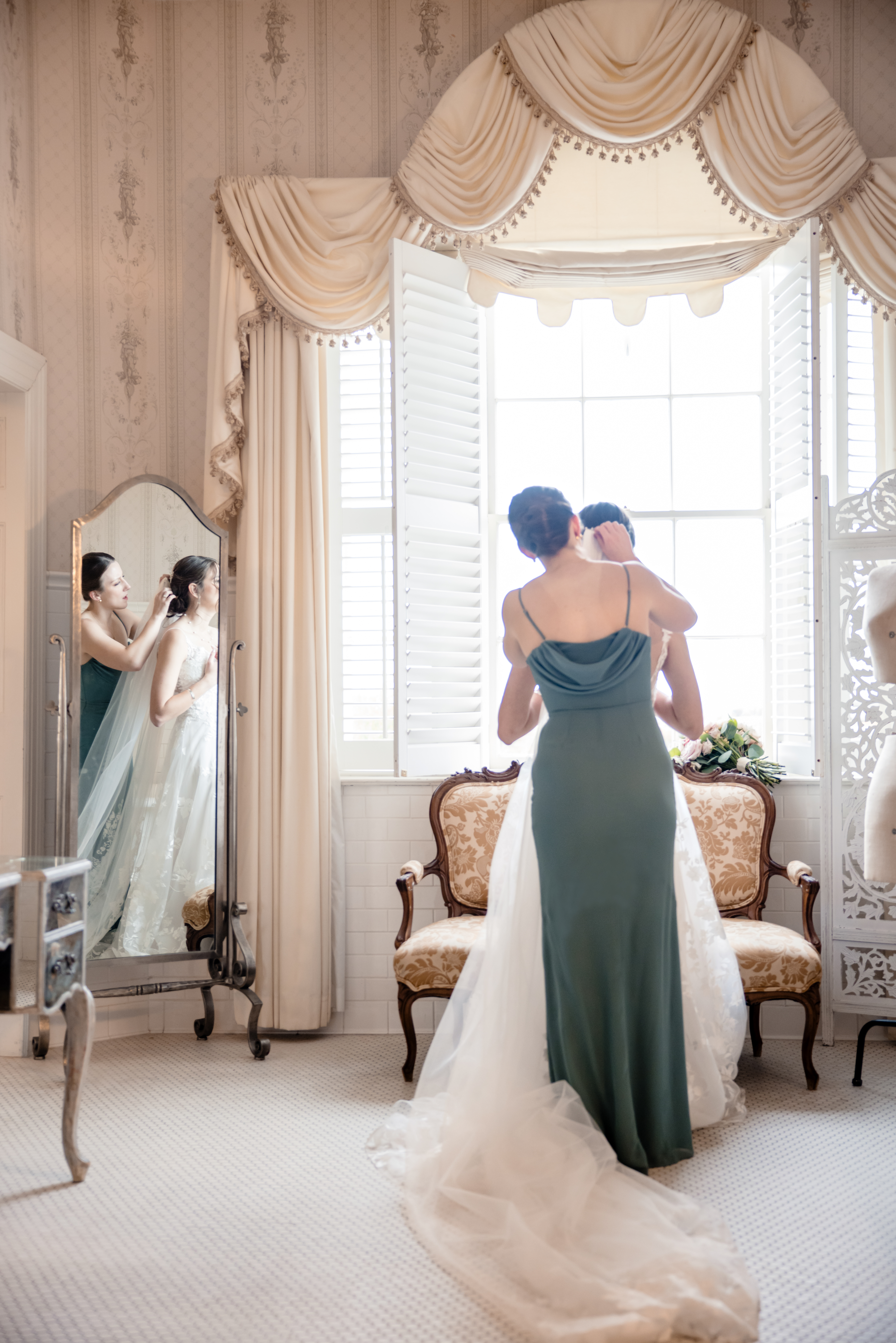 a bridesmaid helping a bride put on her wedding veil in a dressing room