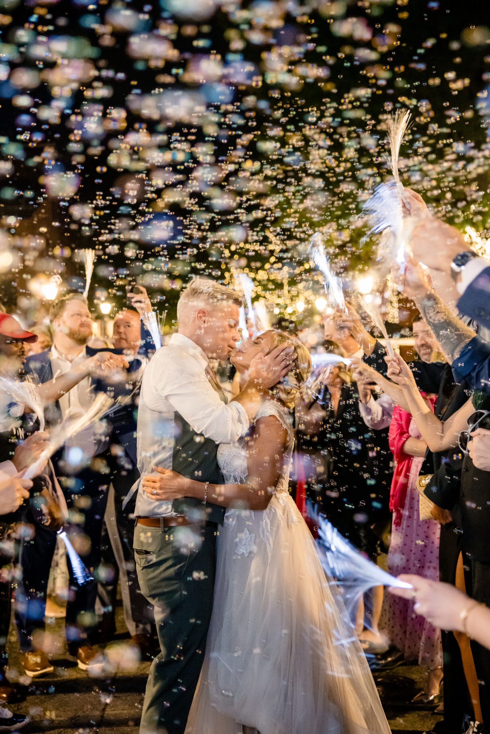 a bride and groom embracing each other and leaning in to kiss one another as their guests wave lights and blow bubbles 