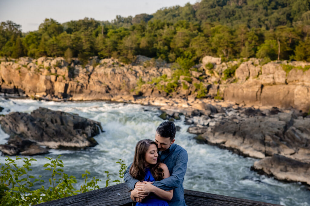 A person standing behind their partner with their arms around their shoulders as they stand in front of a rushing river. 