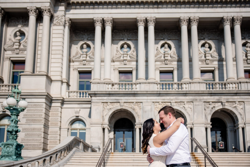 A couple with their arms around each other smiling as they lean in to kiss in front of the Library of Congress 