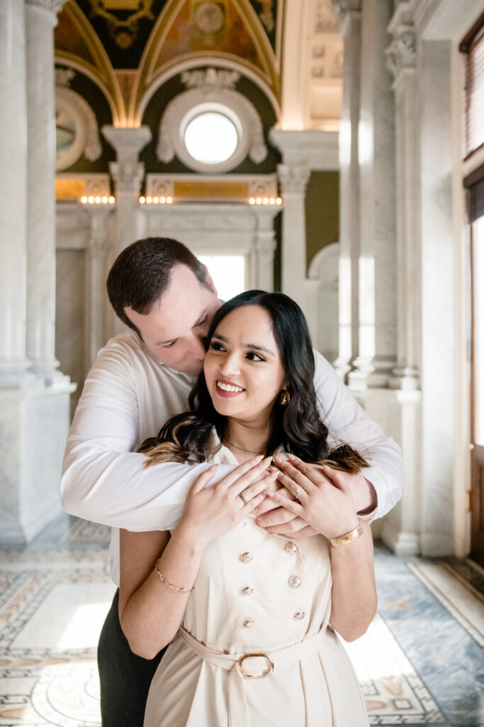 A person standing behind their partner, hugging them from behind and kissing their cheek 