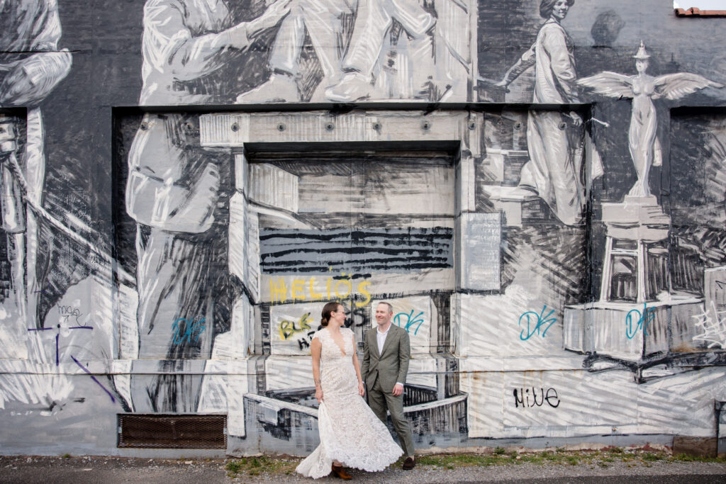 A couple holding hands and laughing together in front of a building with a large mural on it 