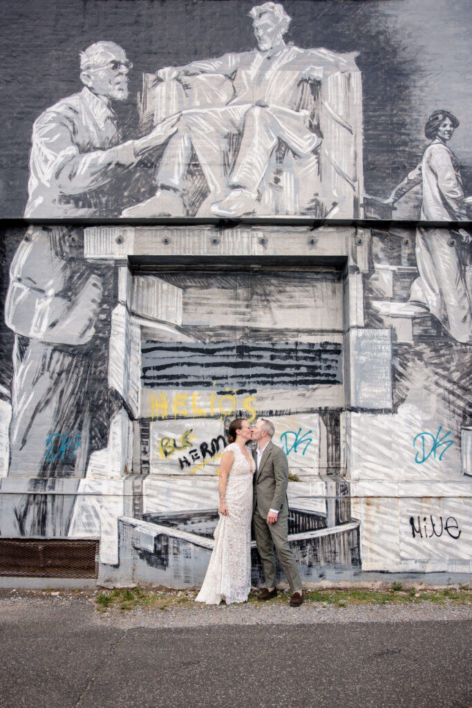 A couple kissing in front of a large black and white mural painted on the side of a building 