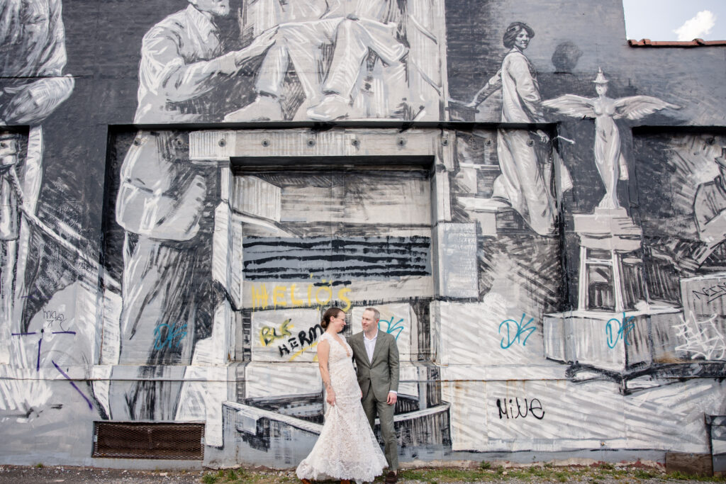 A couple standing side by side with their arms around each other in front of a black and white mural 
