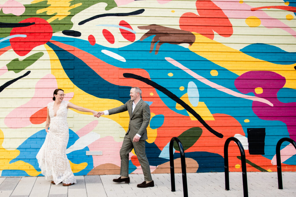 A couple reaching out to hold hands as they walk in front of a colorful mural