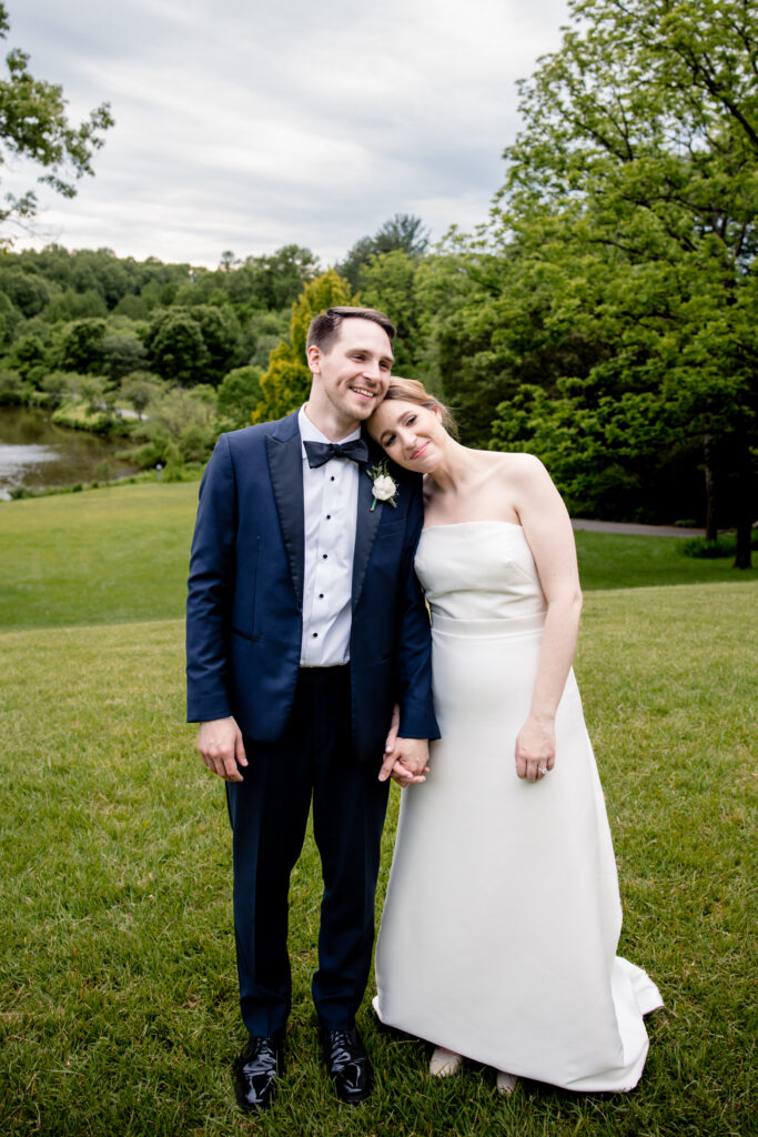 Newlyweds standing in a field holding hands while one rests their head on the other's shoulder 